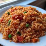Close-up of a serving of fluffy spanish rice mixed with bright green peas and chunks of tomato on a white plate.