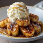 A close-up of warm, caramelized bananas foster topped with a scoop of vanilla ice cream and caramel drizzle.