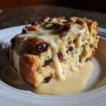 A close-up of a moist slice of bread pudding studded with raisins, generously covered in creamy vanilla sauce.