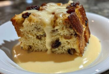 A close-up of a serving of amazing bread pudding topped with raisins and drenched in vanilla cream sauce.