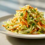 A close-up of a refreshing mound of cabbage salad mixed with bright orange shredded carrots and green parsley.
