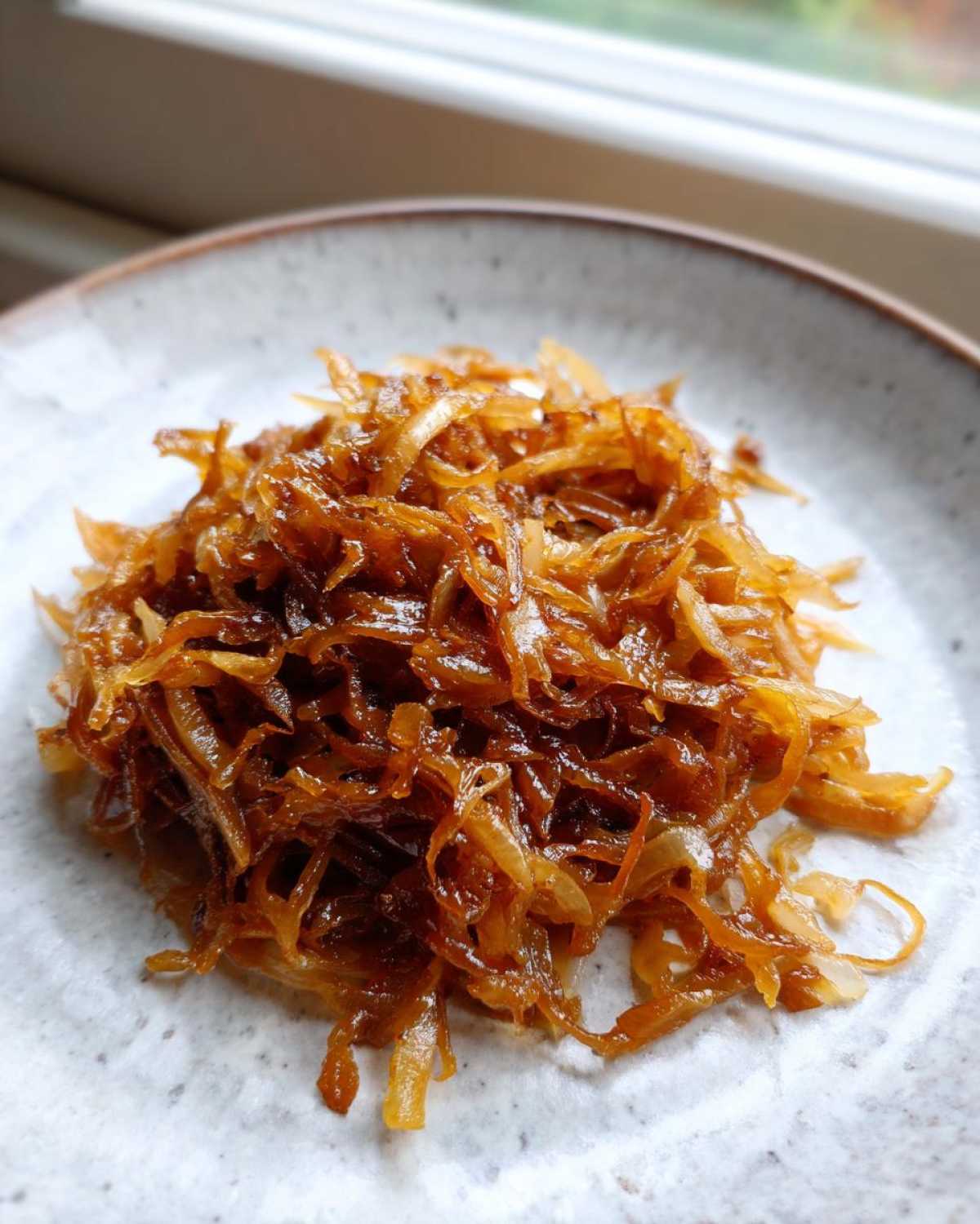 A close-up of a mound of deeply browned, glossy caramelized onions served on a light, textured ceramic plate.