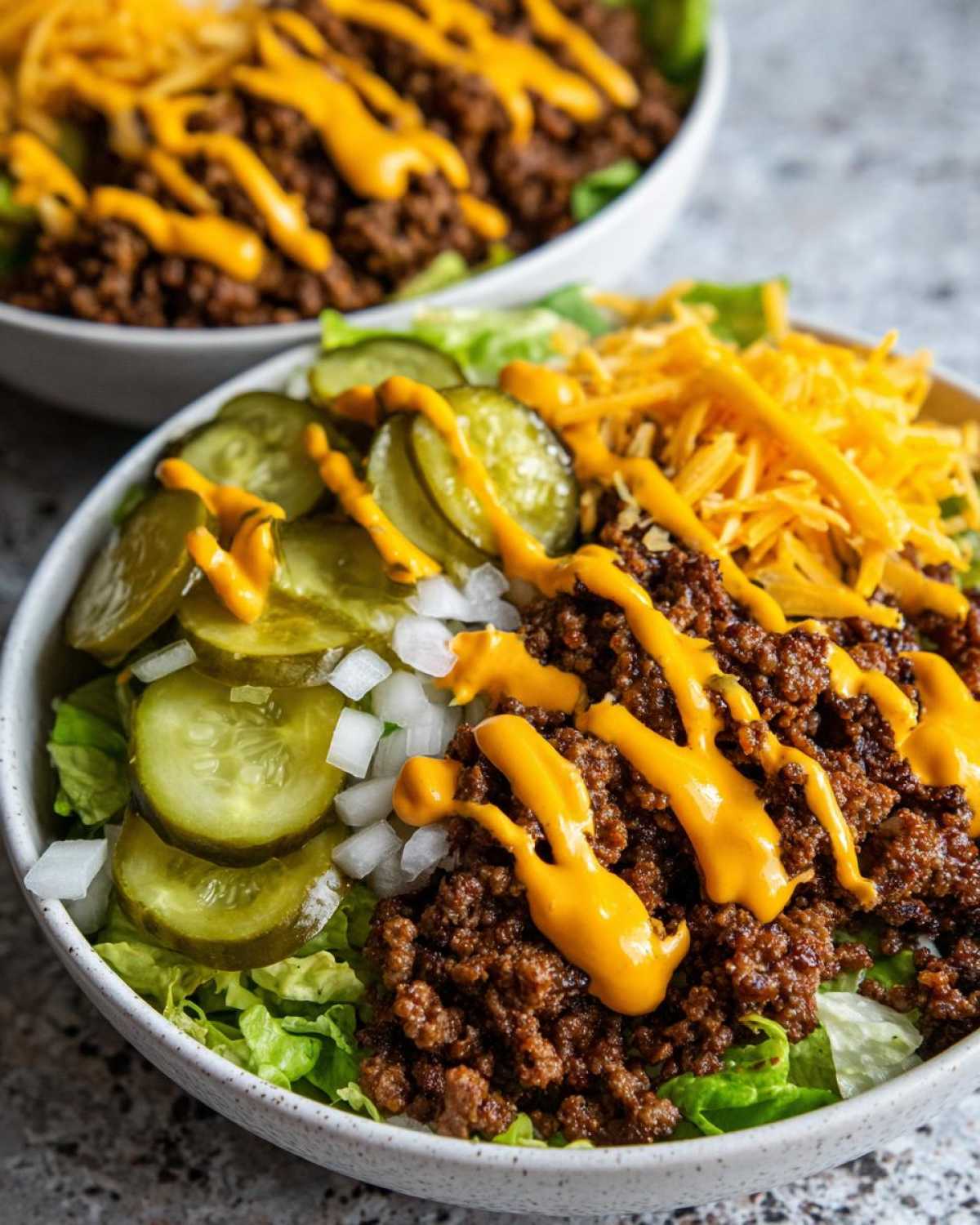 Close-up of a cheeseburger bowl featuring seasoned ground beef, lettuce, pickles, onions, shredded cheese, and a drizzle of sauce.