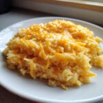 A serving of creamy cheesy rice topped with melted shredded cheddar cheese on a white plate.
