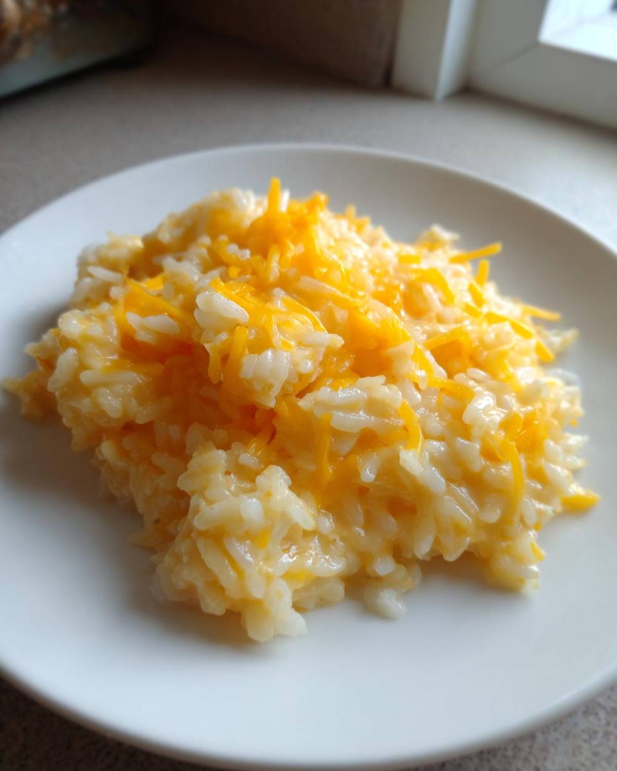 A close-up view of a serving of amazing cheesy rice topped with melted shredded cheddar cheese on a white plate.