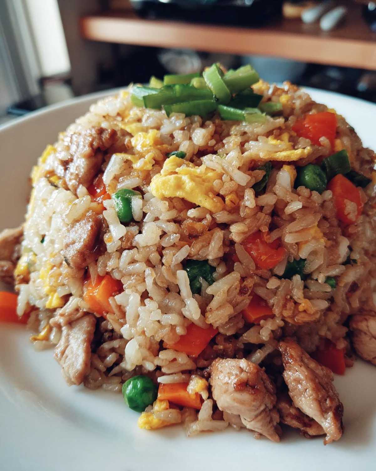 A mound of freshly made chicken fried rice featuring pieces of chicken, carrots, peas, and scrambled egg, topped with green onions.
