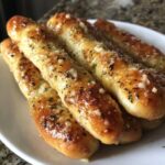 A close-up stack of golden brown, glazed breadsticks topped with melted garlic butter and dried parsley.