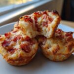 Three golden-brown Pizza Muffins stacked on a white plate, one broken open showing melted cheese and pepperoni filling.