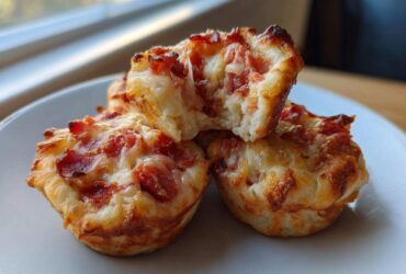 Three golden-brown Pizza Muffins stacked on a white plate, one broken open showing melted cheese and pepperoni filling.