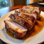Four thick slices of perfectly cooked pork loin roast resting in savory juices on a white plate.