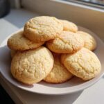 A stack of freshly baked, golden soft sugar cookies with a slightly crackled top, sitting on a light plate.
