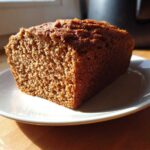 Close-up of a thick slice of dark, moist whole wheat bread resting on a white plate, lit by sunlight.