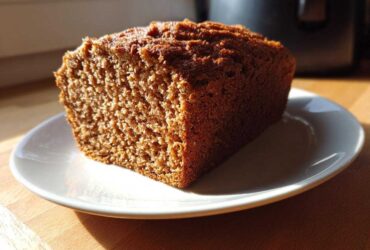 Close-up of a thick slice of dark, moist whole wheat bread resting on a white plate, lit by sunlight.
