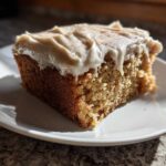 A close-up of a moist slice of applesauce cake topped with creamy frosting and a sprinkle of cinnamon.
