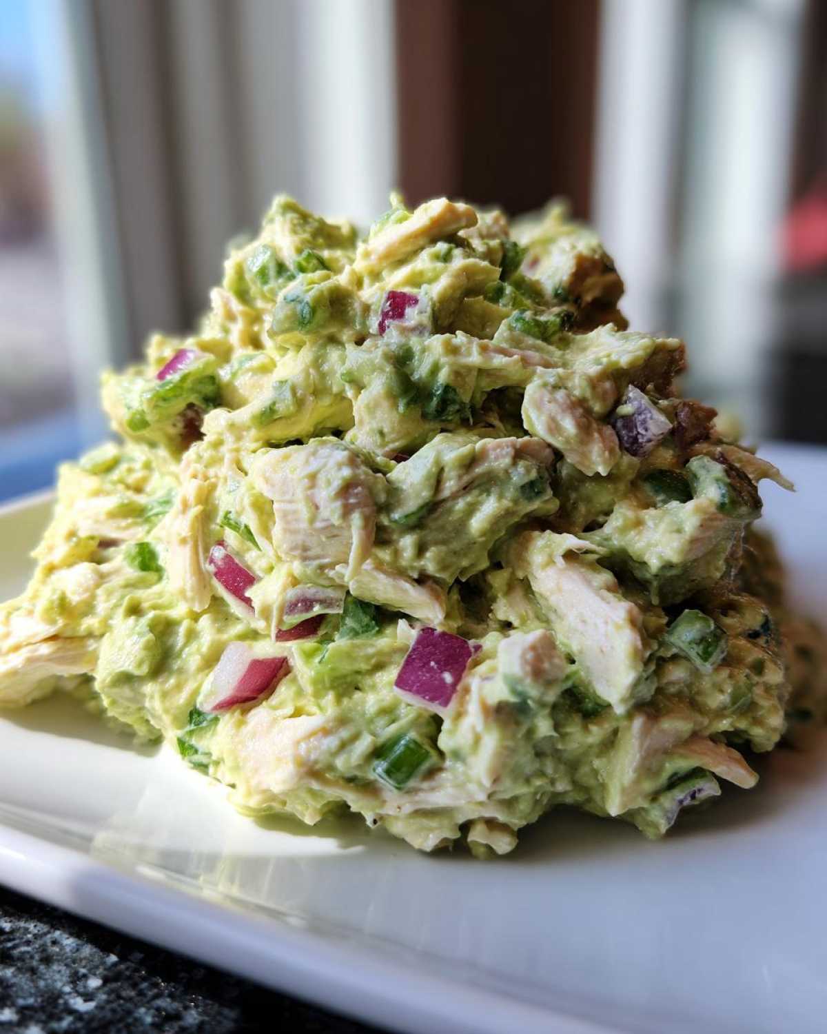 A mound of creamy avocado chicken salad mixed with chunks of chicken and diced red onion, served on a white plate.