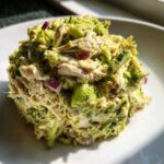 A close-up mound of creamy avocado chicken salad featuring shredded chicken, chunks of avocado, and diced red onion on a white plate.