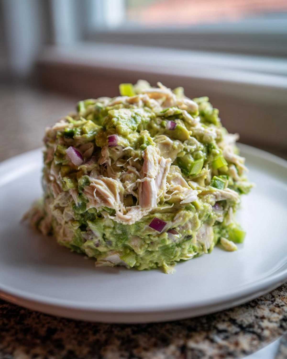 A perfectly molded portion of creamy avocado chicken salad featuring shredded chicken and visible chunks of red onion on a white plate.