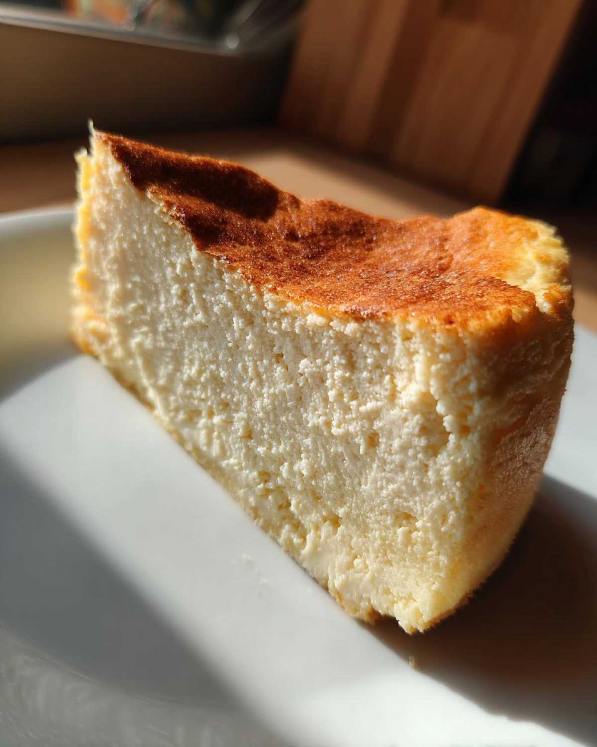 A thick slice of baked Protein Cheesecake with a creamy interior and a deeply browned top, sitting on a white plate.