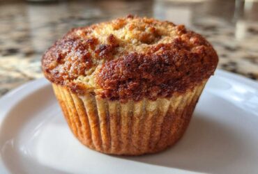 Close-up of a perfectly baked banana protein muffin with a deeply browned, slightly craggy top, sitting on a white dish.