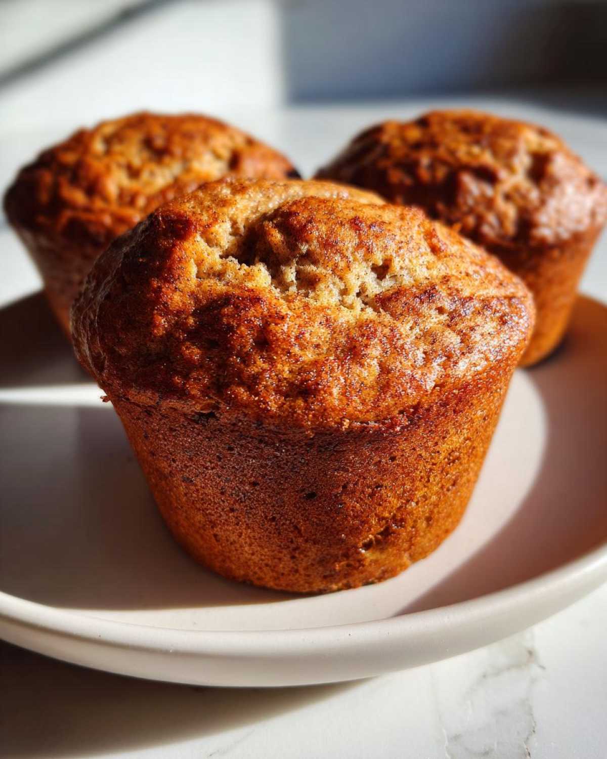 Close-up of three golden brown banana protein muffins sitting on a light-colored plate.