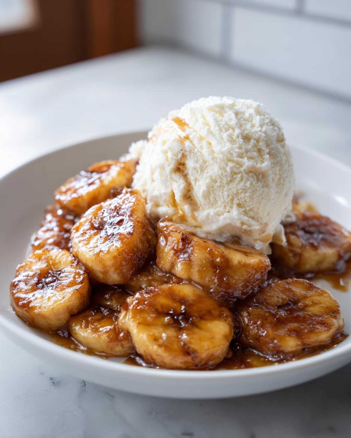 Close-up of caramelized bananas foster topped with a scoop of vanilla ice cream in a white bowl.