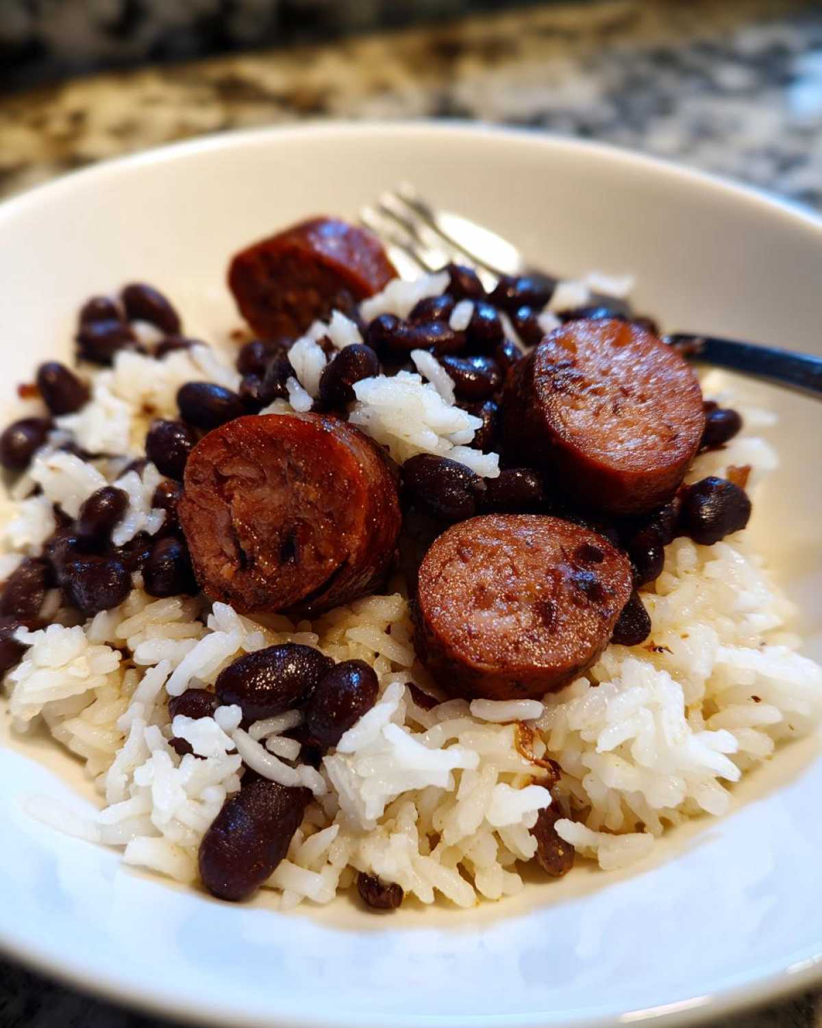 Close-up of a white bowl filled with white rice, black beans and rice, topped with slices of smoked sausage.