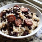 A close-up of a white bowl filled with white rice, black beans and rice, topped with slices of smoked sausage.