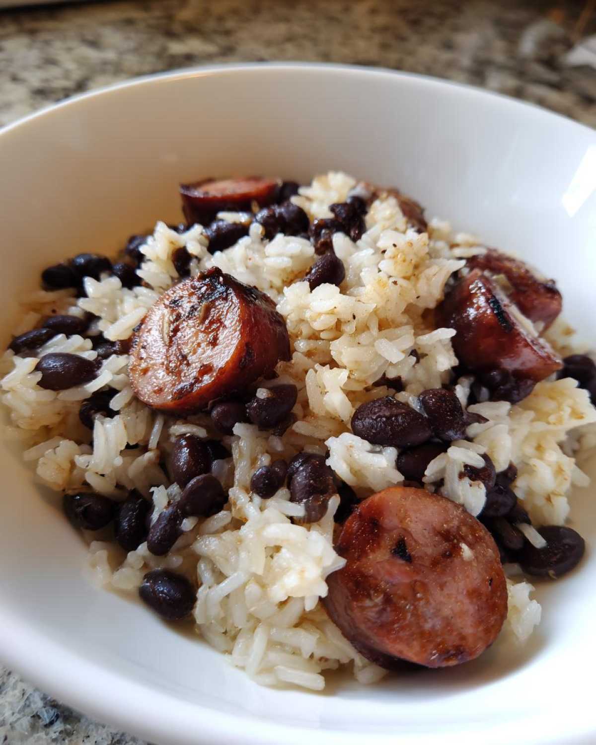 Close-up of a white bowl filled with seasoned rice, black beans and rice, and slices of grilled sausage.