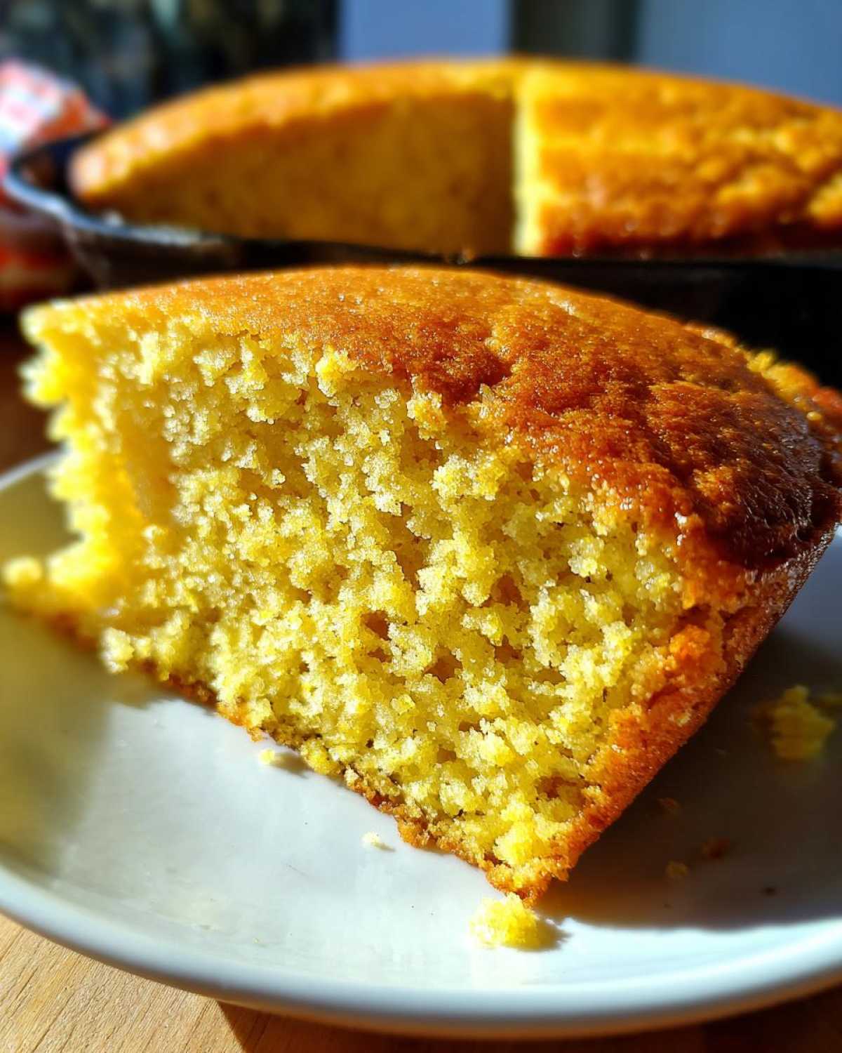 Close-up of a fluffy, golden slice of buttermilk cornbread on a white plate, with the rest of the cornbread visible in a skillet background.