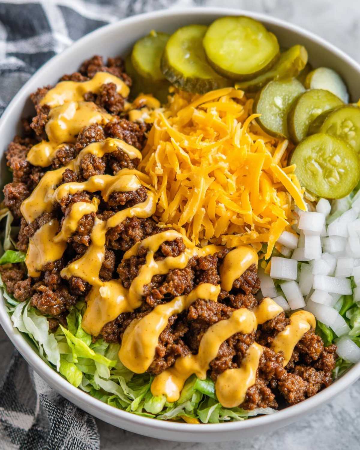 A white bowl filled with a cheeseburger bowl featuring seasoned ground beef, shredded cheese, pickles, onions and sauce over lettuce.