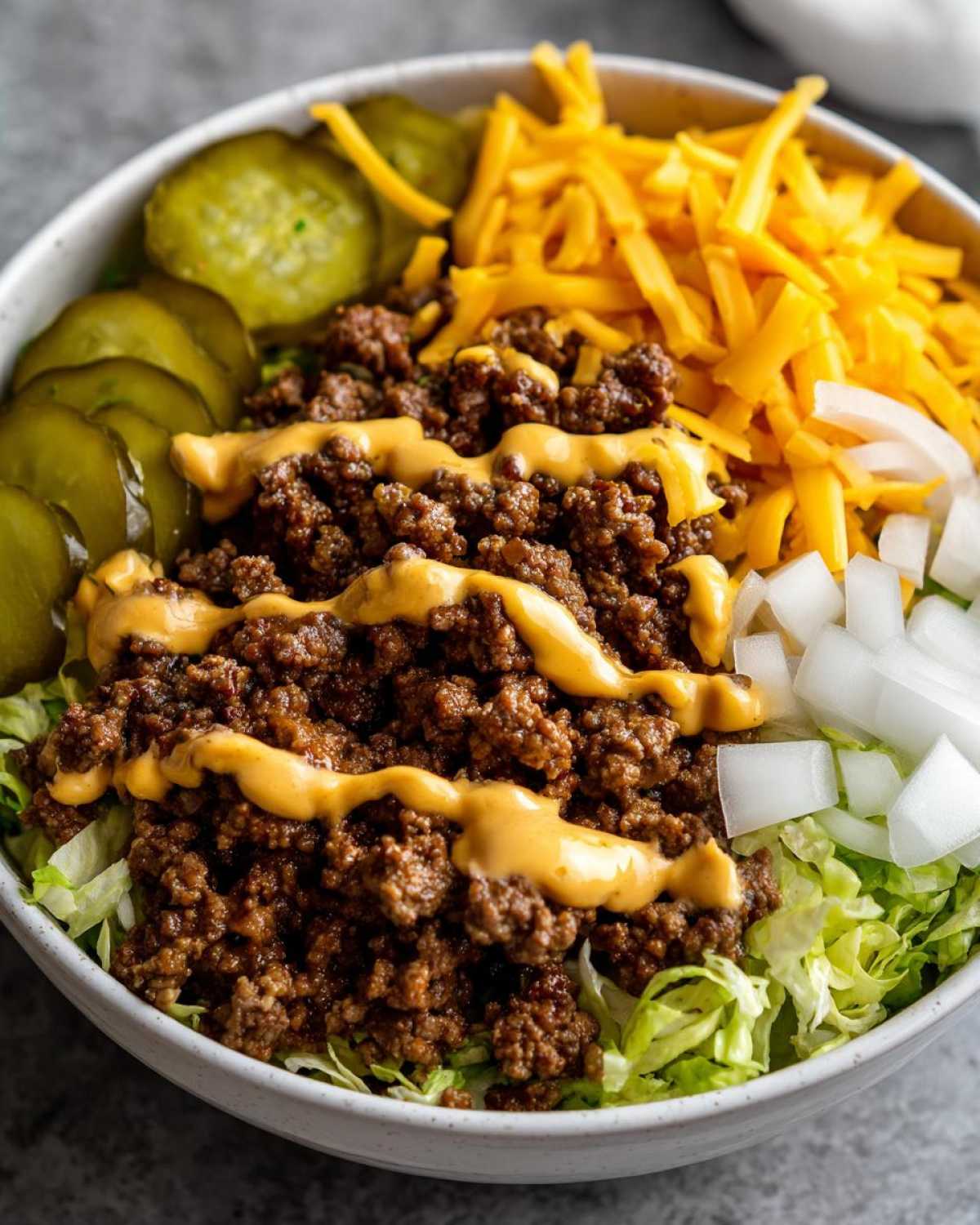 A close-up of a delicious cheeseburger bowl featuring seasoned ground beef, shredded cheddar, pickles, and sauce over lettuce.