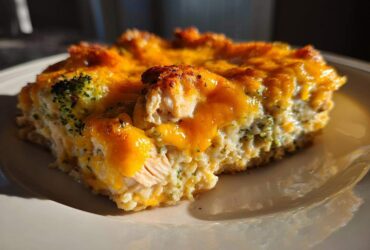 A close-up slice of cheesy chicken and rice casserole topped with melted, browned cheddar cheese and visible broccoli florets.