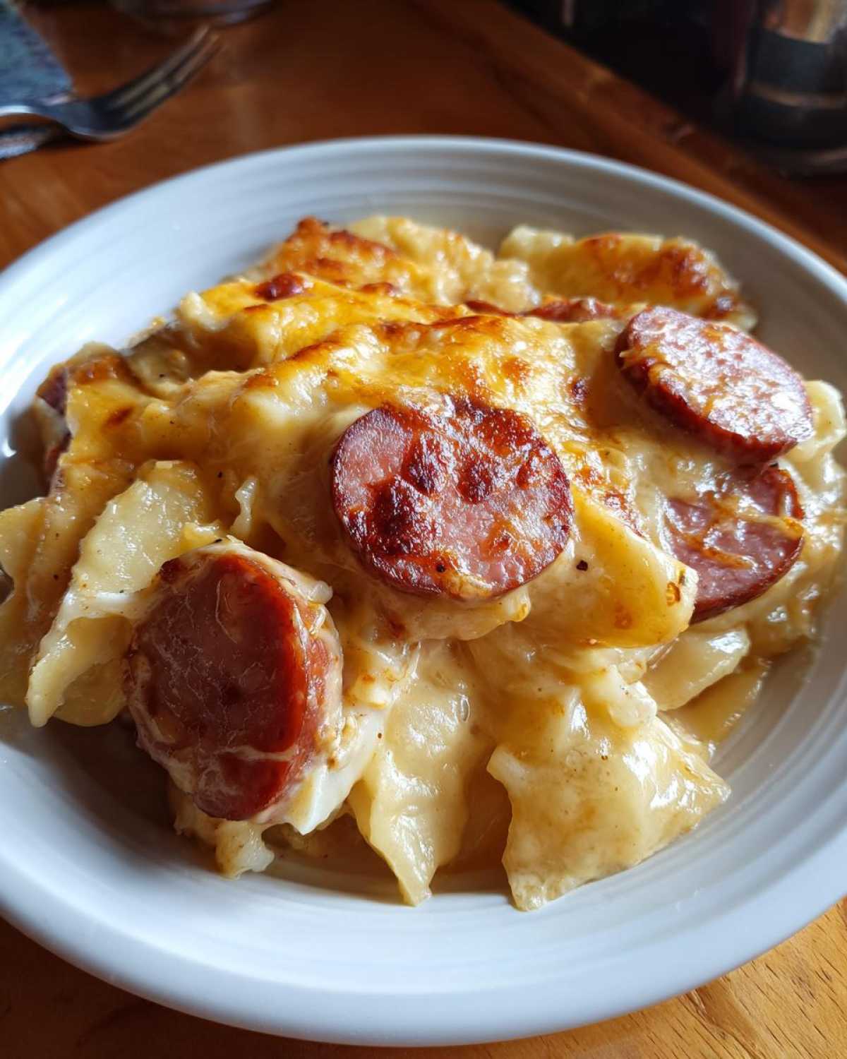 A close-up of a serving of pierogi casserole topped with melted, browned cheese and slices of smoked sausage.