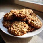 A stack of freshly baked, chewy oatmeal cookie piled high on a white plate, illuminated by natural light.