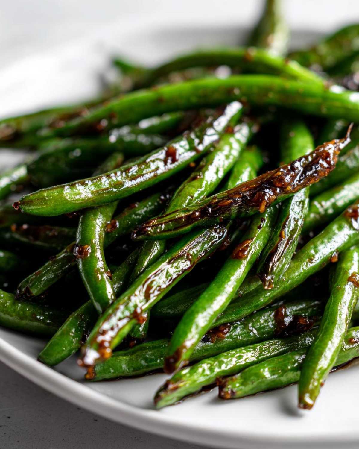 Close-up of vibrant green beans coated in a glossy, dark chinese green beans with garlic sauce, served on a white plate.