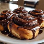 Four freshly baked chocolate cinnamon rolls covered in a thick, glossy chocolate glaze, sitting on a white plate.