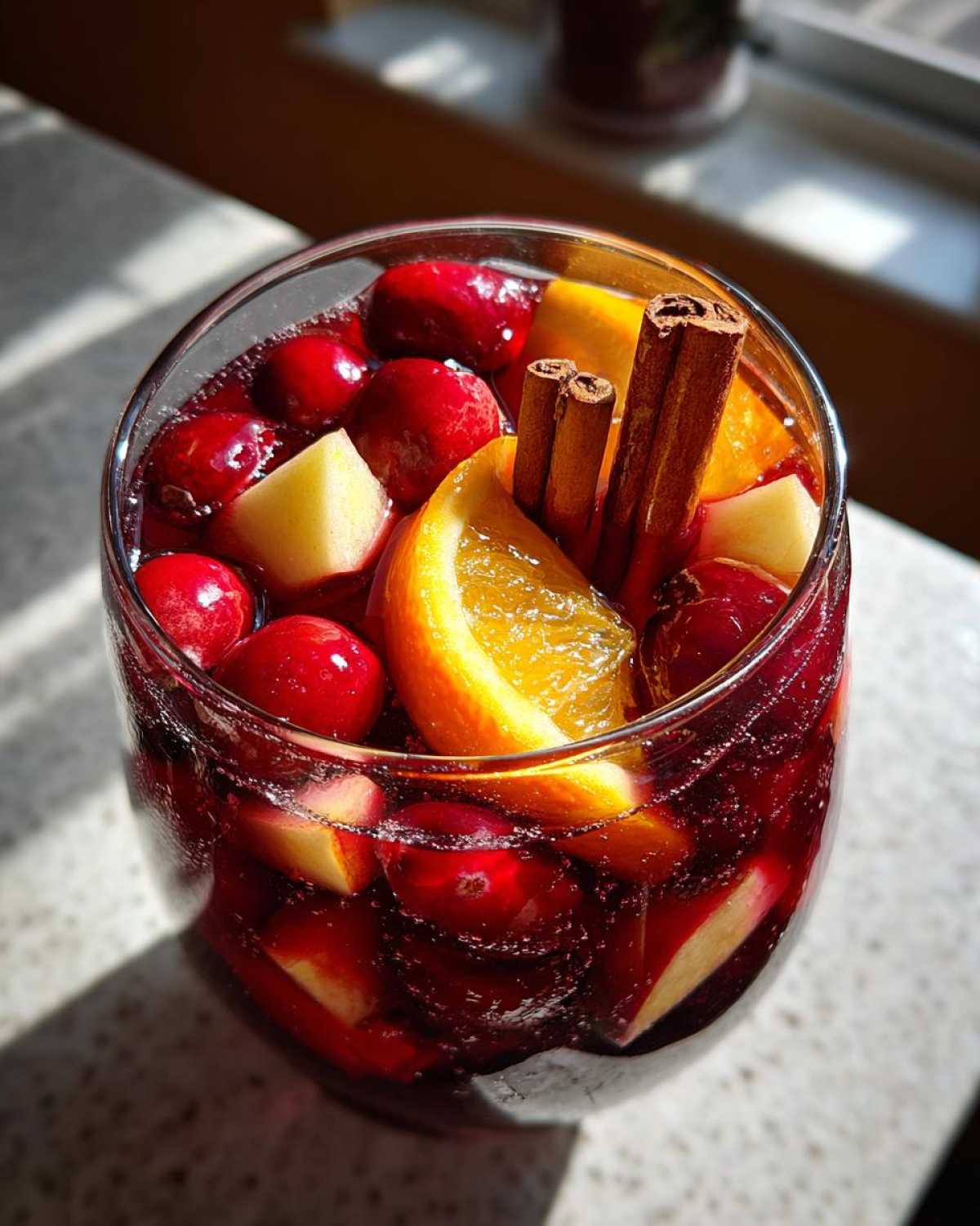 Close-up of a glass filled with vibrant red christmas sangria, garnished with whole cranberries, apple chunks, orange slices, and cinnamon sticks.