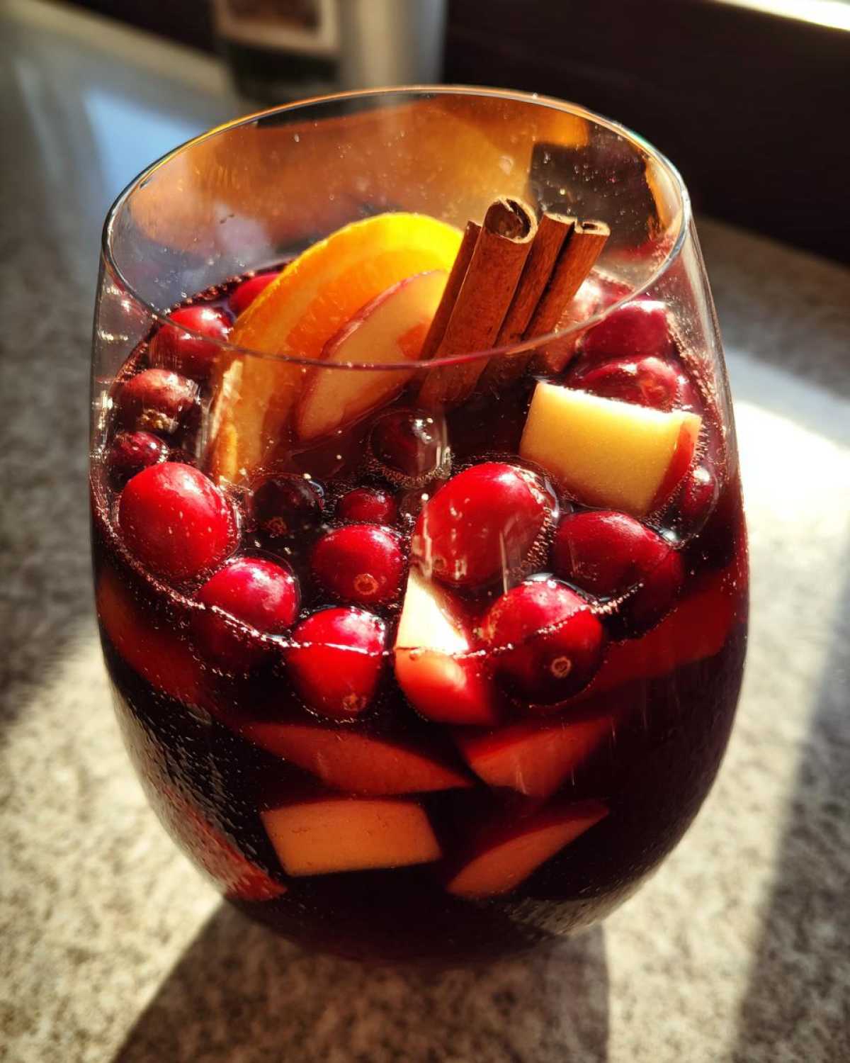A glass filled with rich red christmas sangria, garnished with cranberries, apple slices, orange, and cinnamon sticks.