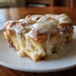A close-up of a gooey slice of cinnamon roll casserole topped with thick white icing.
