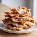 A tall stack of crispy, fried buñuelos generously coated in cinnamon sugar, served on a white plate.