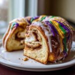 A slice cut from a cinnamon swirl king cake, topped with purple, green, and gold icing.