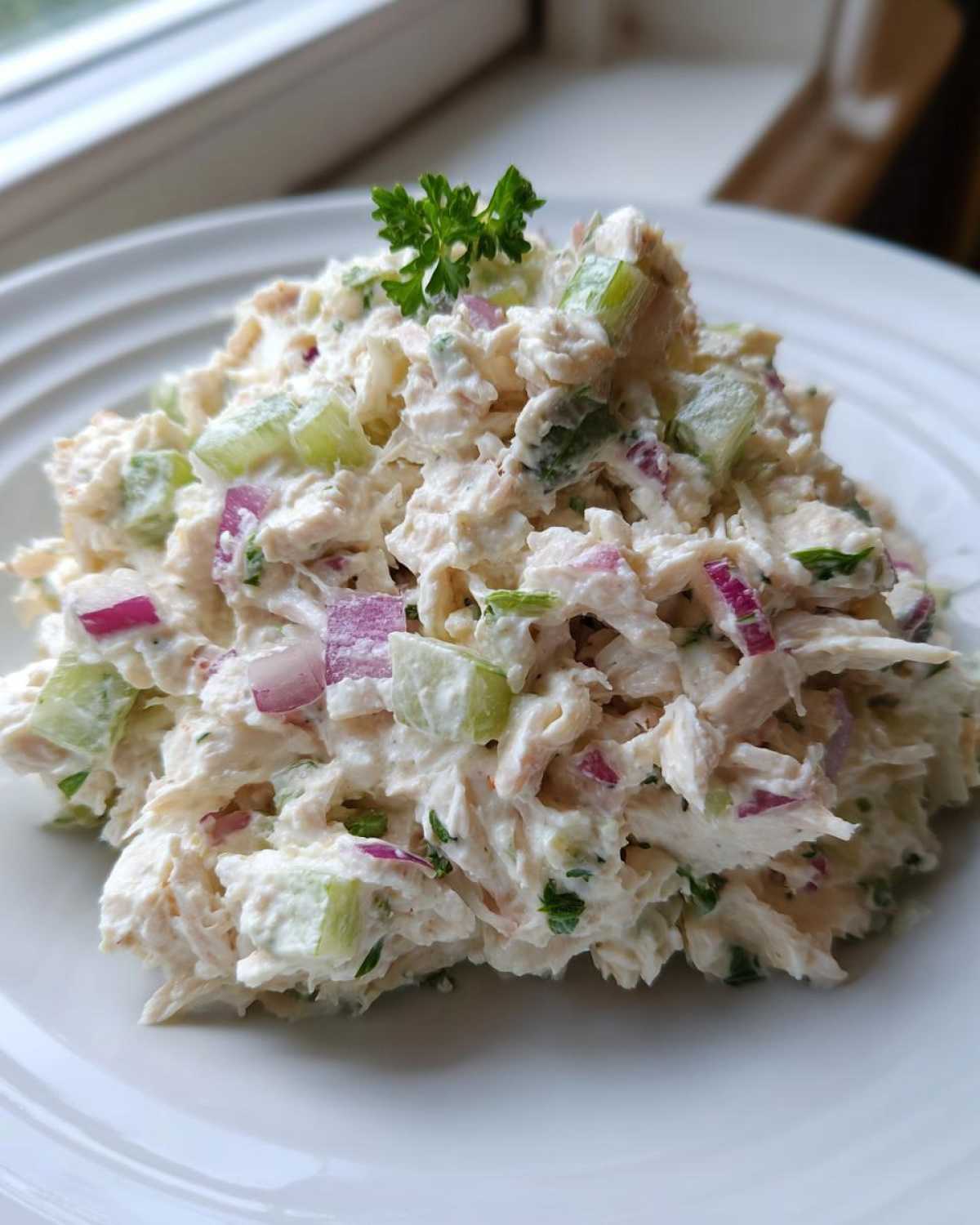 A mound of creamy cottage cheese chicken salad mixed with red onion and celery, garnished with parsley on a white plate.