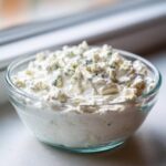 Close-up of a glass bowl filled with creamy blue cheese dip, topped generously with blue cheese crumbles.