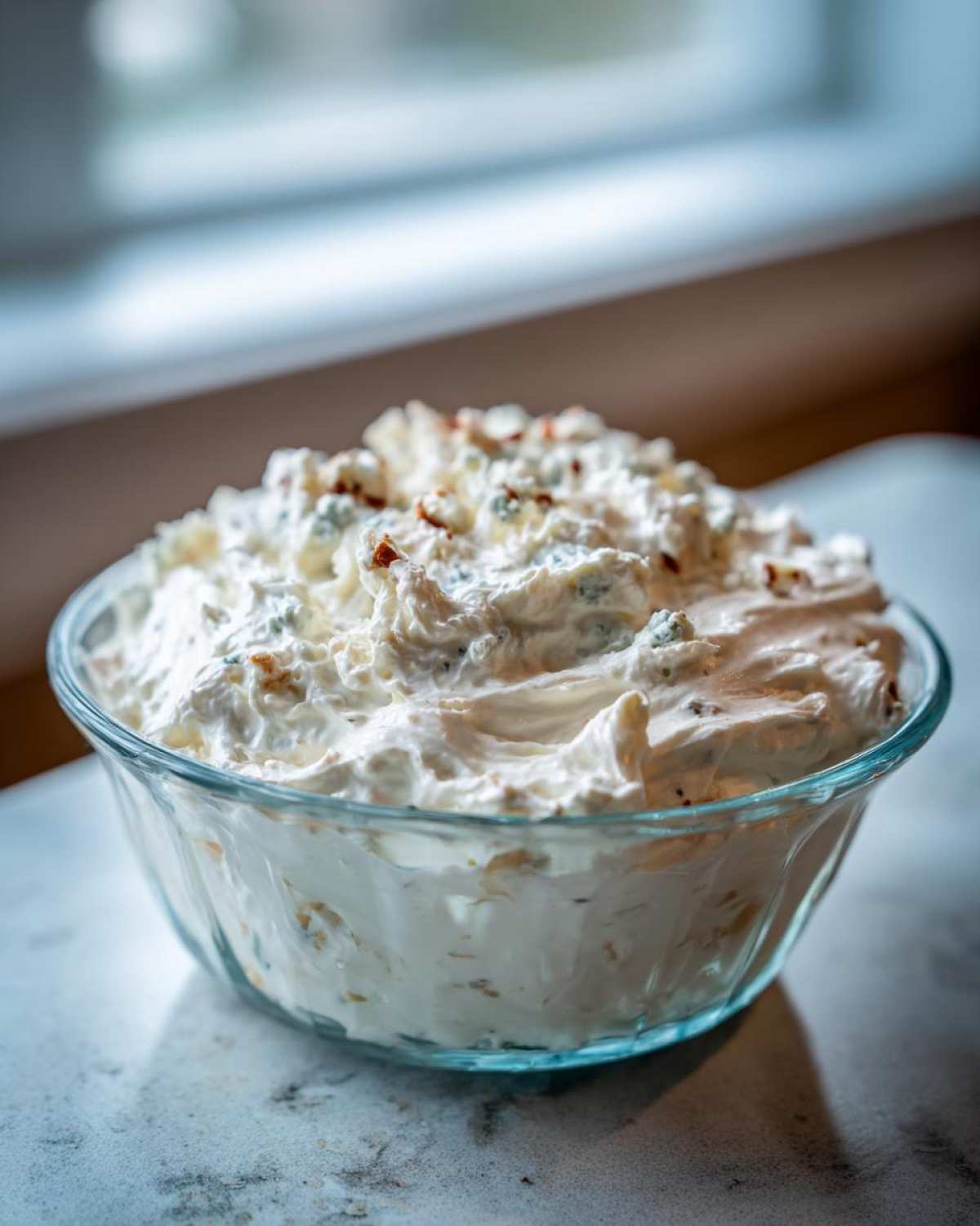 A close-up of a large glass bowl filled with fluffy, creamy blue cheese dip topped with visible blue cheese crumbles.