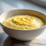 A close-up of creamy, yellow cheese grits served in a white bowl, lightly seasoned on top.
