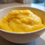 Close-up of creamy, bright yellow cheese grits topped with black pepper, served in a white bowl.