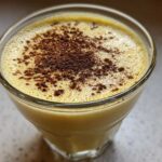 Close-up of a glass filled with creamy, yellow eggnog topped with dusted cocoa or nutmeg.