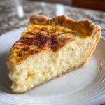 A close-up of a slice of creamy egg custard pie dusted with cinnamon on a white plate.