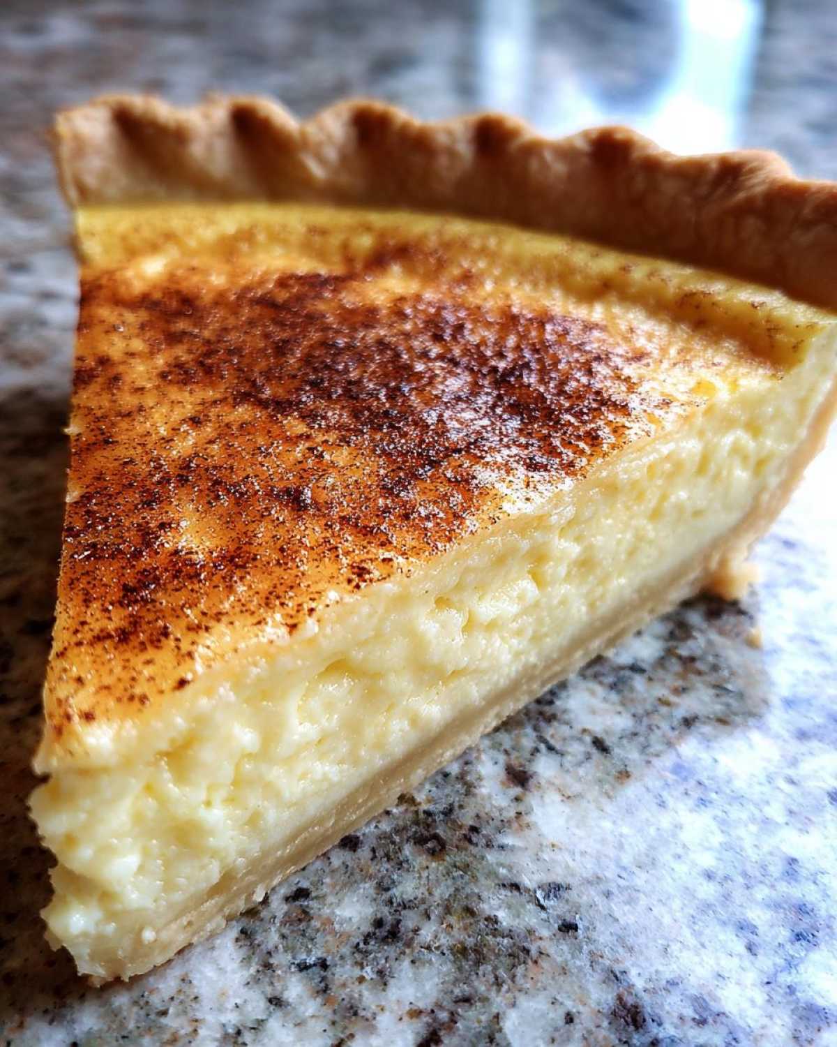 Close-up of a slice of creamy egg custard pie with a golden crust and sprinkled nutmeg on top.