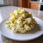 A generous serving of creamy egg salad recipe, featuring chunky white egg pieces and green herbs, piled high on a white plate.